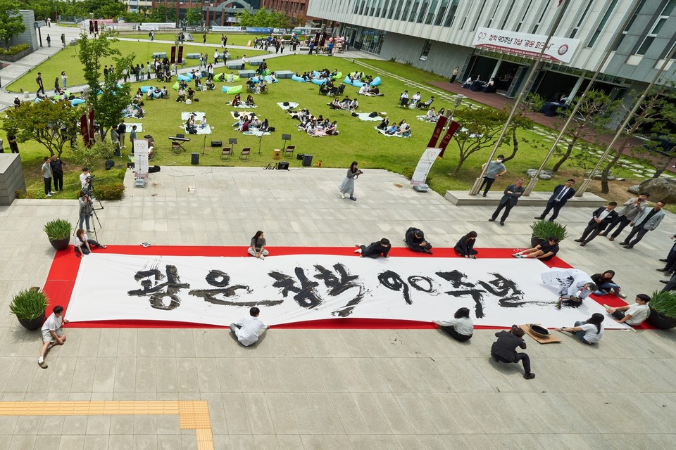 울산 리홍재 서예가가 5월 20일 진행된 ‘광운 창학 90주년’ 기념행사에서 광운대 화도관 앞에 준비된 가로 15미터, 세로 3미터 규모의 대형 천에 ‘광운 창학 90주년’이라는 글씨를 쓰는 퍼포먼스를 선보이고 있다. (사진=광운대)